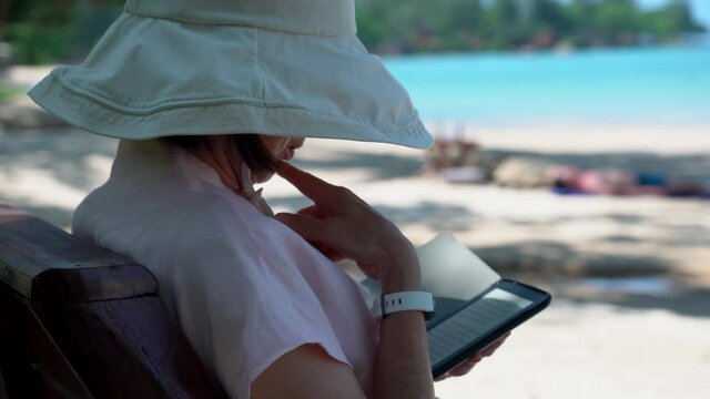 senior woman reading ebook from e reader on beach on summer vacation
