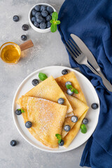 Homemade crepes served with fresh blueberries and powdered sugar on a white plate on a gray concrete background. Food for Maslenitsa. Copy space.