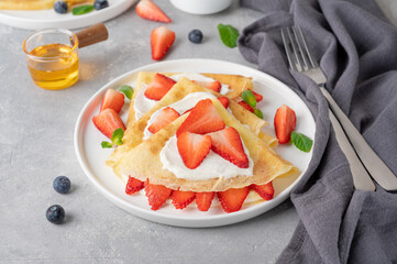 Crepes or thin pancakes with cream cheese, fresh strawberries and honey on a white plate on a gray concrete background. Copy space.