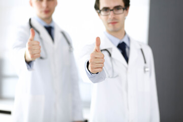 Two male doctors standing as a team and showing thumbs up as a symbol of the best service for patients in the clinic. Medicine and health care