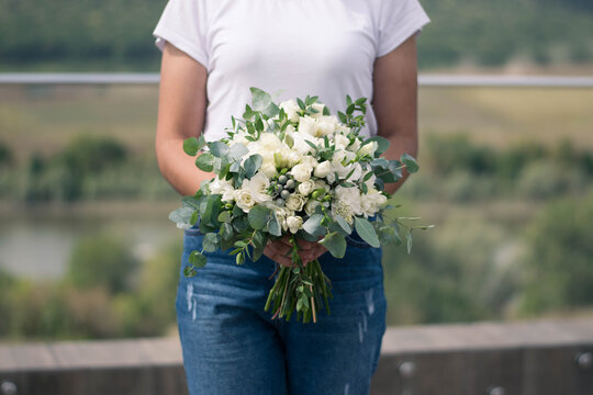 Florist hands holding modern bridal bouquet of white flowers and greenery. Wedding bouquet composed of roses, freesia, lisianthus, brunia and eucalyptus.