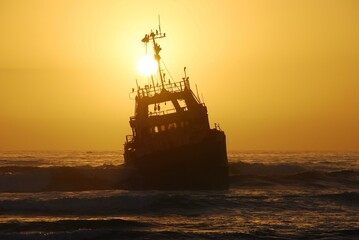 Ship wreck sitting on a reef at sunset on the coast of Namibia, the sun is behind the ship, yellow...