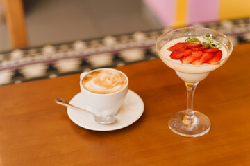 Creamy dessert with strawberries in glass, cappuccino on wooden table. Appetizing dessert and latte on the background of the restaurant