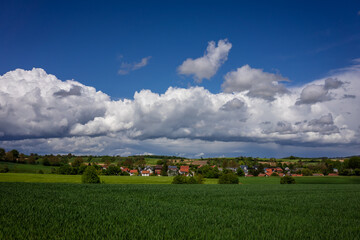 Obraz premium Schöne, grüne Frühlingslandschaft im Kraichgau, Deutschland, mit Getreidefeld im Vordergrund und einem Dorf mit bewölktem Himmel im Hintergrund.