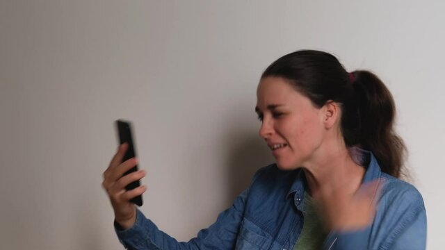 Young Caucasian European woman on white background talks on video link using smartphone and looks at phone screen smiles and blows kiss. 4K footage. Conversation over Internet with loved one.