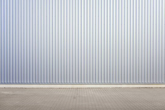 Exterior wall of warehouse made of aluminum sheet and paved road in outdoor area as background image. Texture of a wall made of silver corrugated metal sh