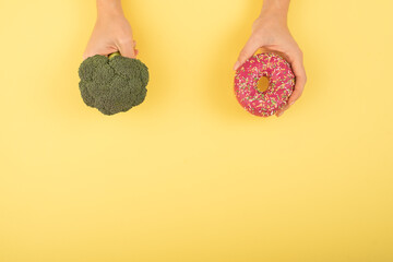 Comparison of eating habits. Woman holding broccoli and donut on a yellow background. Cope space.
