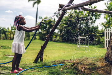 Portrait of happy smiling little child african american girl playing and watering garden grass with rubber strap and sunny summer in garden at home