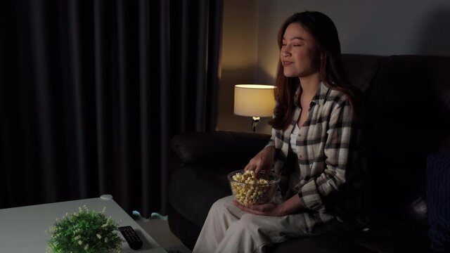 funny young woman watching TV and eating popcorn on sofa at night