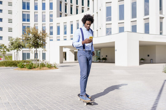 Black businessman on a skateboard looking at his smartphone outdoors. - Powered by Adobe