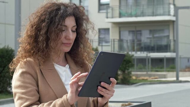 Curly-haired woman middle-aged walks along a deserted city street. She communicates with someone through a tablet.