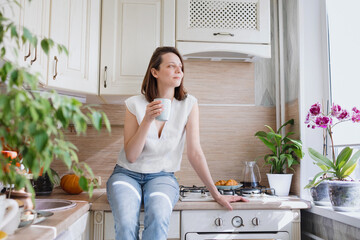 a European woman sits on the table in her kitchen and drinks coffee, breakfast in the kitchen...