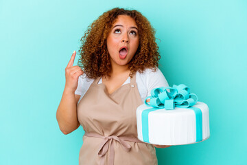 Young pastry latin woman holding a cake isolated on blue background pointing upside with opened mouth.
