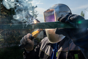 Fototapeta premium The welder welds a new metal frame for the fence around the house in backdrop trees, village