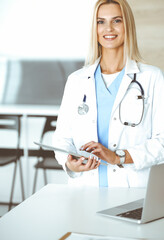 Woman-doctor at work in clinic excited and happy of her profession. Blond female physician is smiling while using tablet computer. Medicine concept