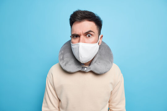 Indoor shot of serious attentive man wears protective mask against coronavirus inflated neck pillow for comfort sleep during travelling in transport raises eyebrow looks attentively at camera