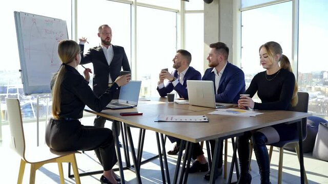 Office room with business partners. Group of young entrepreneurs sitting at the table and listen to a cheerful businessman.