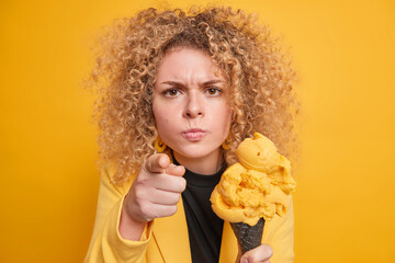 Young curly haired woman looks angrily and points directly at camera blames you holds delicious ice cream eats yummy summer dessert dressed in formal clothes isolated over yellow background.