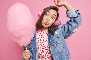 Surprised Asian woman tilts head keeps lips folded raises arm listens music via stereo headphones holds delicious candy floss wears denim jacket isolated over pink background. Delicious dessert