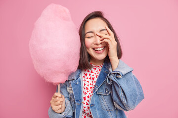 Happy sincere Asian woman has carefree expression hand on face smiles joyfully being in good mood holds sweet candy floss wears stylish denim jacket has free time expresses positive emotions