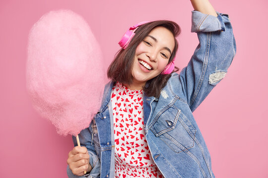 Joyful Teenage Girl Tilts Head Raises Arm Has Fun Listens Music Via Wireless Headphones Dressed In Fashionable Denim Jacket Holds Sugary Cotton Candy Isolated Over Pink Background. People Lifestyle