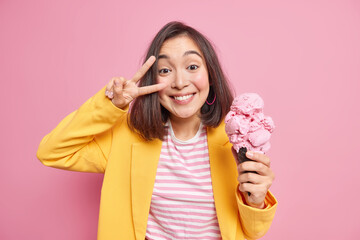 Happy joyful Asian woman makes victory sign over eye smiles toothily holds big cone ice cream in waffle dressed in stylish clothes has fun during hot summer day isolated over pink background.
