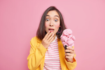 Photo of surprised young Asian woman with dark hair stares shocked dressed in striped t shirt and yellow jacket holds yummy ice cream isolated over pink background. People and emotions concept