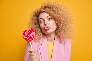 Lovely affectionate woman with curly hair keeps lips folded holds delicious candy on stick has...