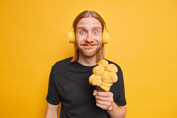 Horizontal shot of positive bearded man with long ginger hair smeared face with ice cream holds big yellow gelato dressed casually has fun enjoys summer holidays listens music via headphones