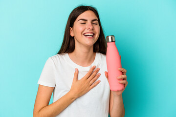 Young caucasian woman holding water bottle isolated on blue background laughs out loudly keeping...