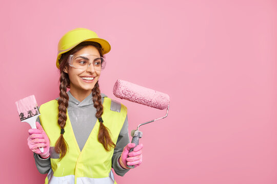 Positive busy female builder involved in renovation and repair of building holds roller and paint brush follows strict safety regulations wears protective equipment has leadership skills. Copy space