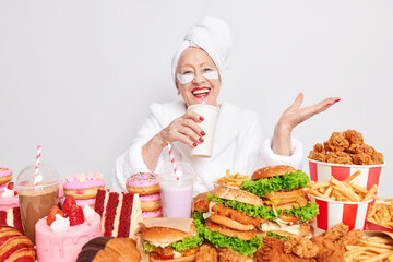 Positive carefree old woman wears shower towel on head beauty patches under eyes red lipstick cares about herself drinks soft drink eats junk food has unhealthy nutrition no restrictions and diets