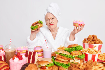 Displeased mature woman holds delicious doughnut and burger has unhealthy eating habits eats harmful food cannot avoid cosuming cheat meal addicted to sugar and fat dressed in domestic robe.