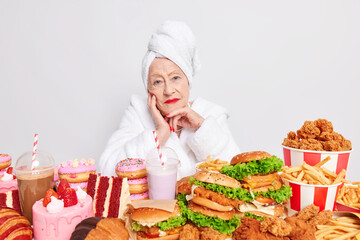 Senior lady in bathrobe has clean wrinkled face sits at table full of appetizing junk food has...