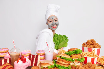 Happy woman stands sideways smiles toothily wears bathrobe and towel on head applies clay facial mask for skin treatment keeps to weight loss diet holds green lettuce stands near variety of junk food