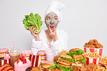 Healthy lifestyle proper nutrition temptation concept. Pensive Asian woman wears beauty face mask and red lipstick holds green lettuce salad chooses between healthy and unhealthy food. Diet failure