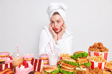 Indoor shot of lovely woman enjoys softness of skin after beauty procedures wears bath towel and white robe poses near table full of junk food being addicted to cheat meal. Overeating concept