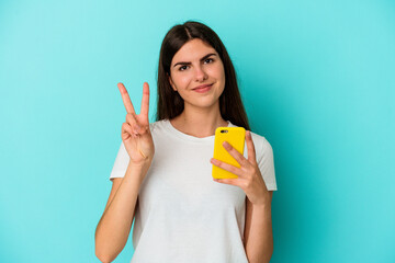 Young caucasian woman holding a mobile phone isolated on blue background showing number two with fingers.