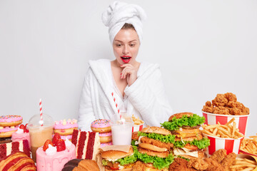 Unhealthy eating concept. Surprised woman has red lips being very hungry looks at table overloaded with hamburgers milkshakes and cakes isolated over white background wears bath towel dressing gown
