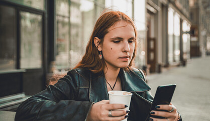 portrait of young teenager redhead girl with long hair with smartphone and cup of hot coffee at city street cafe terrace