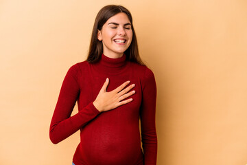 Fototapeta premium Young caucasian pregnant woman isolated on beige background laughs out loudly keeping hand on chest.
