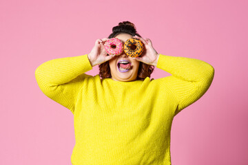 Cheerful plus size model holding donuts against her eyes on pink background