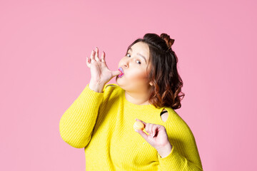 Happy plus size model eating macaroon, fat girl loves sweets, pink studio background