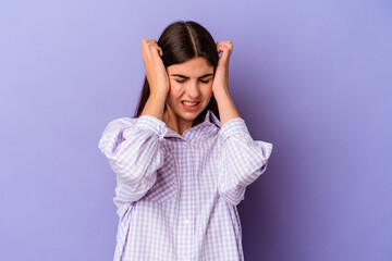 Young caucasian woman isolated on purple background covering ears with hands.