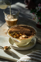 Portion of classic tiramisu in glass bowl, spoon and cup of  coffee with milk on wooden board.