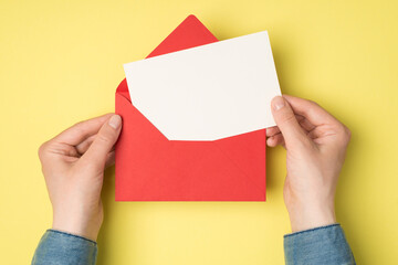 First person top view photo of female hands holding open red envelope with white paper card on isolated yellow background with blank space