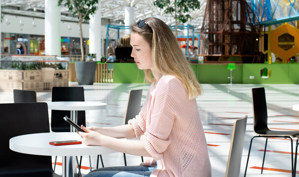 Young woman wearing pink cardigan is using digital tablet sitting at the table in a cafe of a shopping mall or at the airport. Freelance work 
