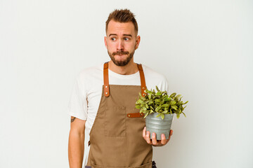 Young gardener tattooed caucasian man holding a plant isolated on white background confused, feels doubtful and unsure.