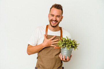 Young gardener tattooed caucasian man holding a plant isolated on white background laughs out...