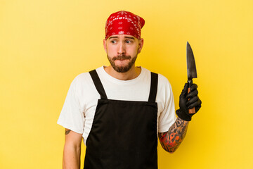 Young tattooed batcher caucasian man holding a knife isolated on yellow background confused, feels doubtful and unsure.